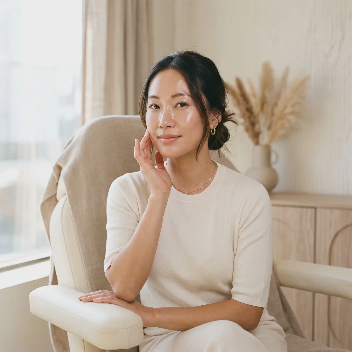 Woman with dewy, healthy-looking skin touching her cheek in a sunlit treatment room at Beautiful Body and Beyond in Corona, CA